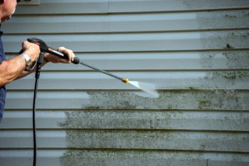 An Unidentified Man Uses A Power Washer To Clean Mold And Grime Off The Siding Of A House