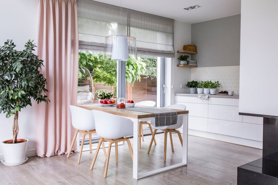 Gray Roman Shades And A Pink Curtain On Big, Glass Windows In A Modern Kitchen And Dining Room Interior With A Wooden Table And White Chairs