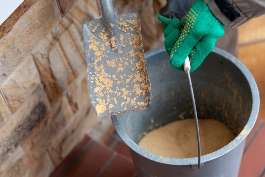 Hands In Green Work Gloves Hold A Plastic Bucket Of Dry Sand And A Small Spatula. Sand Stuck To The Shoulder Blade. Close-Up