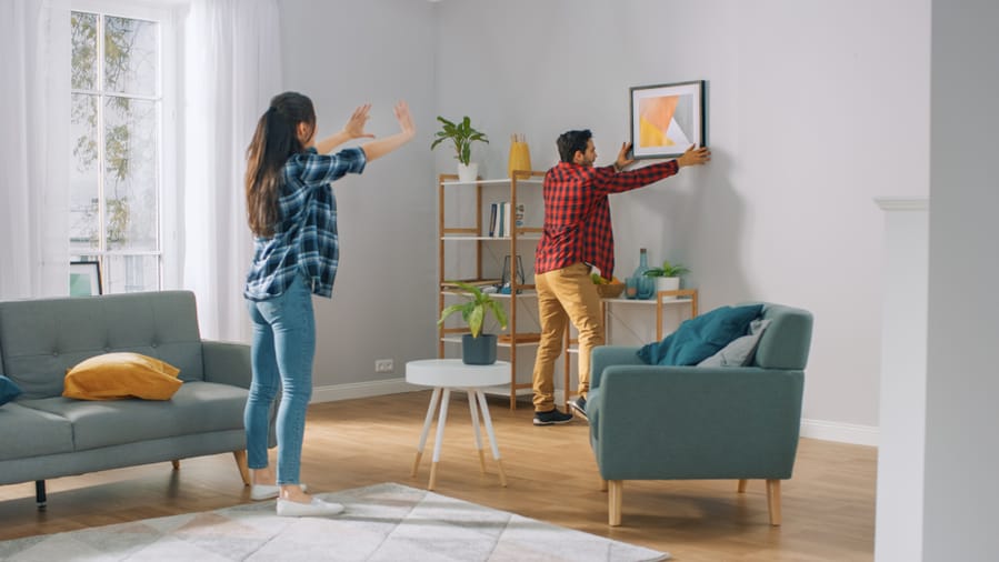 Happy Couple Hanging Picture On The Wall, Boyfriend Moves It, Girlfriend Tells Him When The Frame Is Hanging Straight.