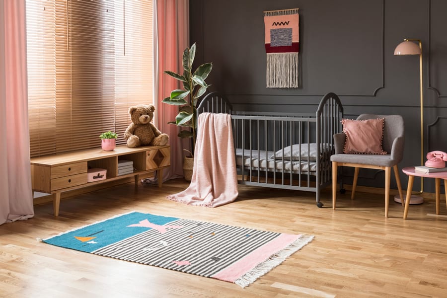 Real Photo Of A Baby Crib Standing Between A Low Cupboard And An Armchair, Lamp And Stool In Child's Room Interior With Wooden Floor And Grey Walls With Molding