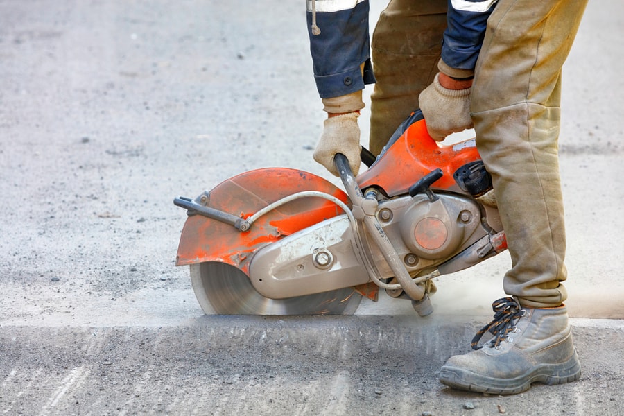 A Worker Mends Part Of The Road, Cuts Out Worn Asphalt In A Cloud Of Dust Using A Portable Concrete Cutter And A Cutting Diamond Blade, Copy Space, Close-Up.