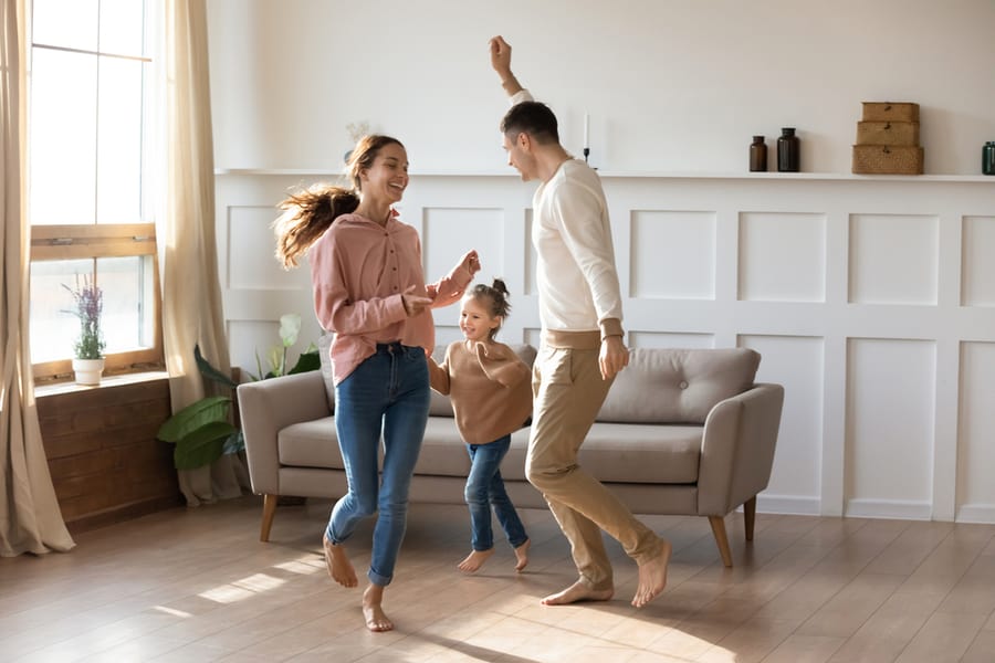 Cheery Couple Dancing With Little Daughter Barefoot On Wooden Laminate Floor With Underfloor Heating System In Modern Warm Living Room.