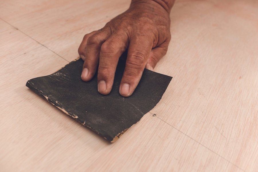 An Older Handyman Using A Piece Of Sandpaper To Smoothen Out The Surface Of A Sheet Of Plywood Prior To Painting.